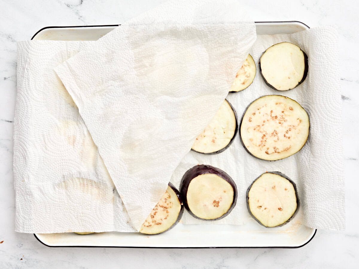 Eggplant slices being dried between two layers of paper towel on a baking sheet.
