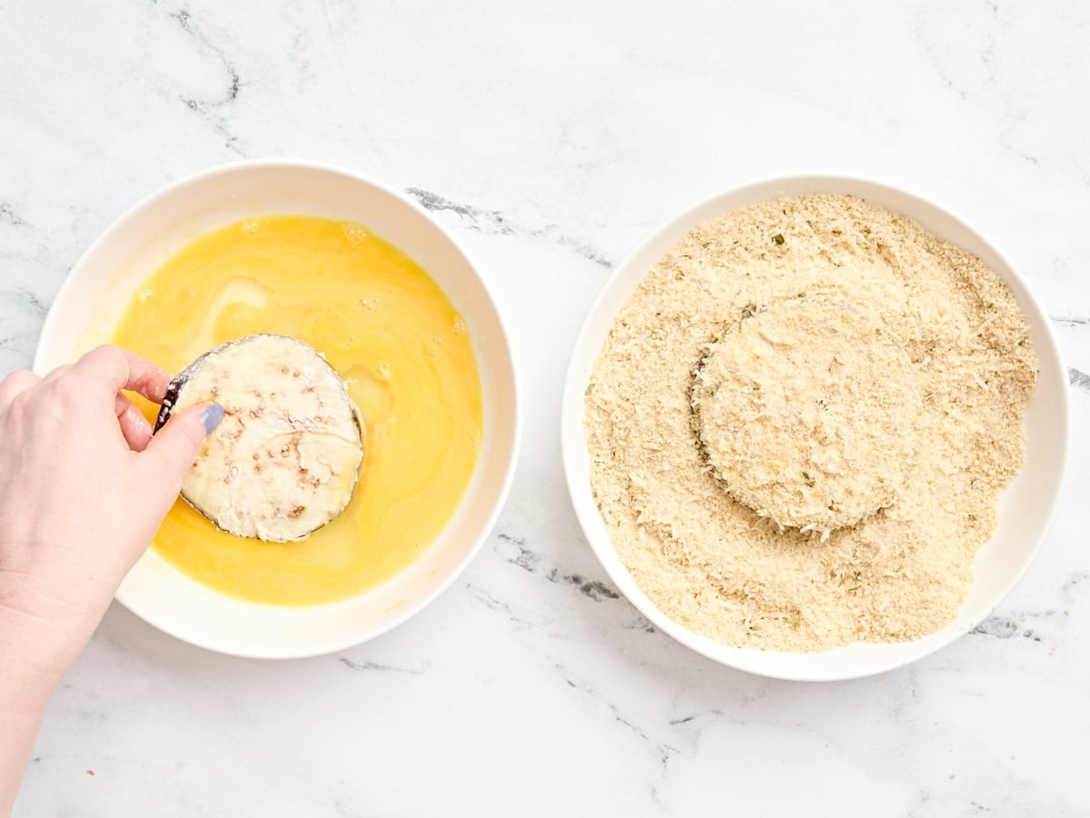 Floured eggplant slices being dipped into beaten egg and then breadcrumbs to make a breadcrumb coating.