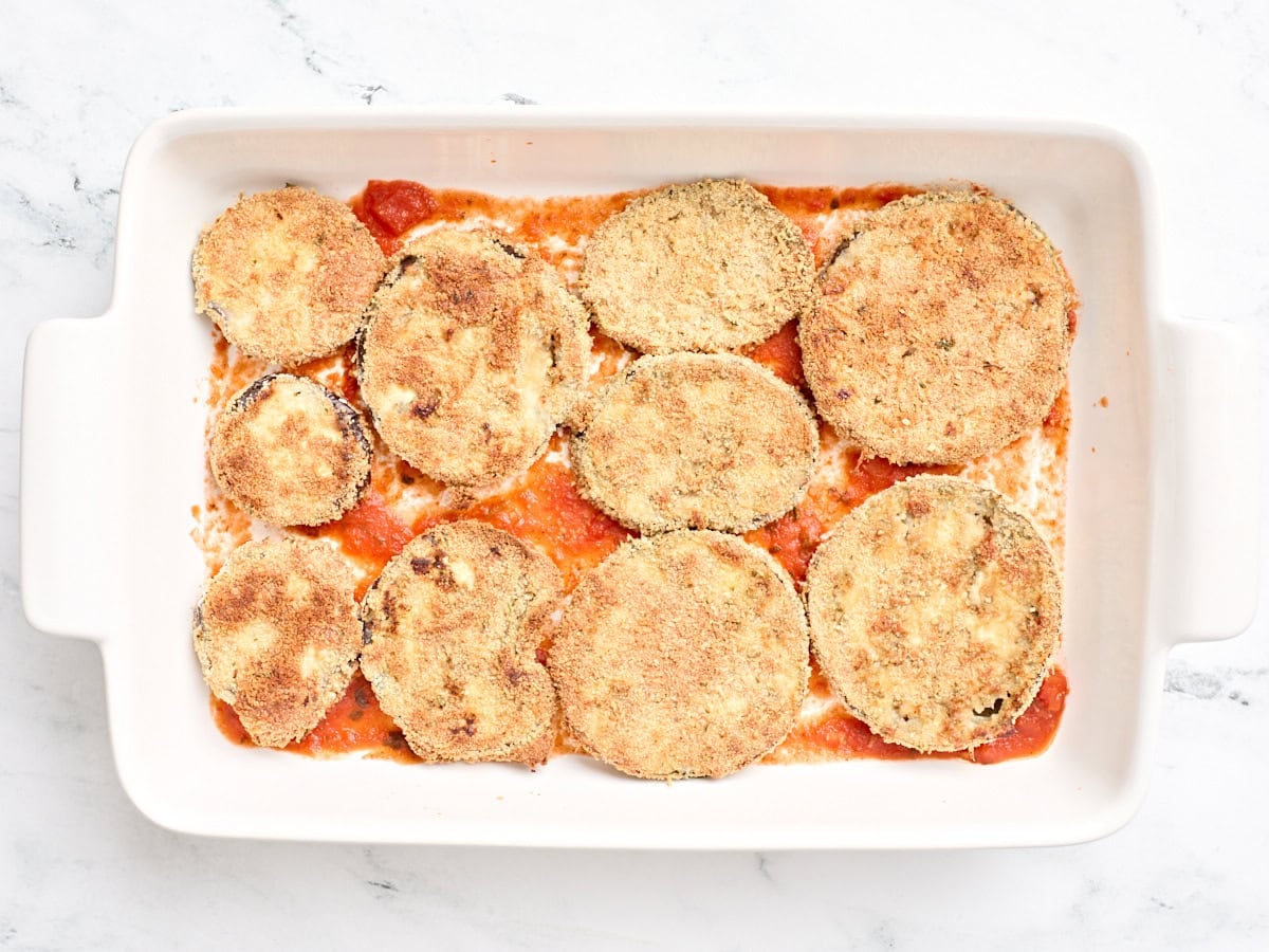 Breaded parmesan slices on a layer of marinara sauce in a baking dish.
