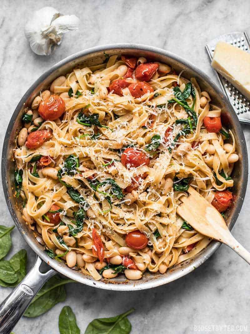 This Tuscan White Bean Pasta is a fast and flavorful dish that is perfect for weeknight dinners. Caramelized garlic, basil, and Parmesan add BIG flavor! Budetbytes.com Overhead view of the skillet full of tuscan white bean pasta with a wooden pasta fork in the side, garlic and Parmesan near the skillet