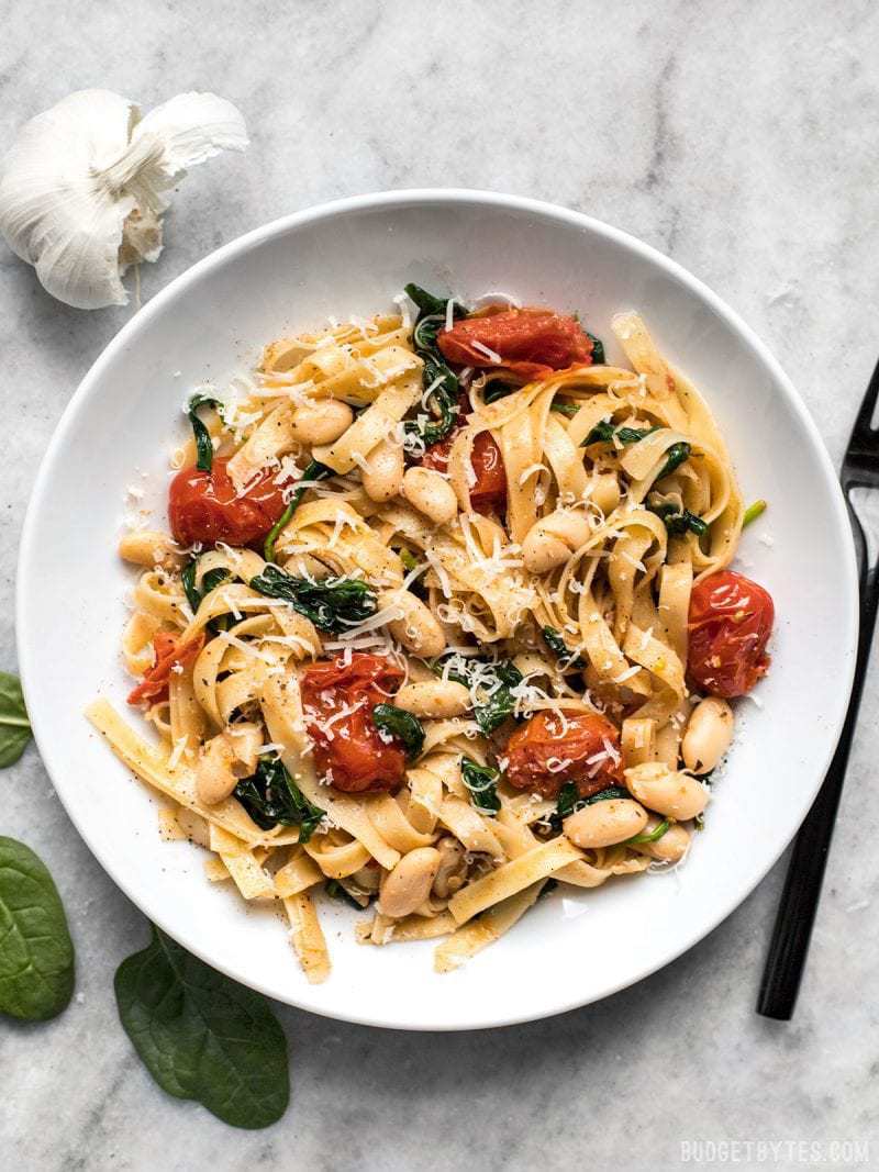 This Tuscan White Bean Pasta is a fast and flavorful dish that is perfect for weeknight dinners. Caramelized garlic, basil, and Parmesan add BIG flavor! Budetbytes.com Overhead view of a large bowl full of Tuscan White Bean Pasta, spinach and garlic on the side