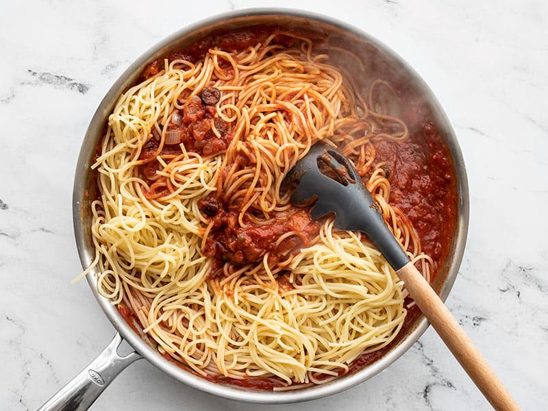Adding cooked spaghetti to puttanesca sauce in the skillet.
