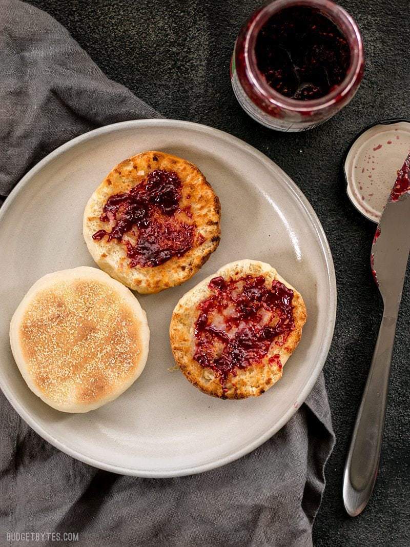 Homemade English muffins are fun to make, delicious, and cost just pennies each. Make this your next weekend project! Step by step photos. BudgetBytes.com Two English muffins on a plate, one sliced open and toasted, smeared with raspberry jam.
