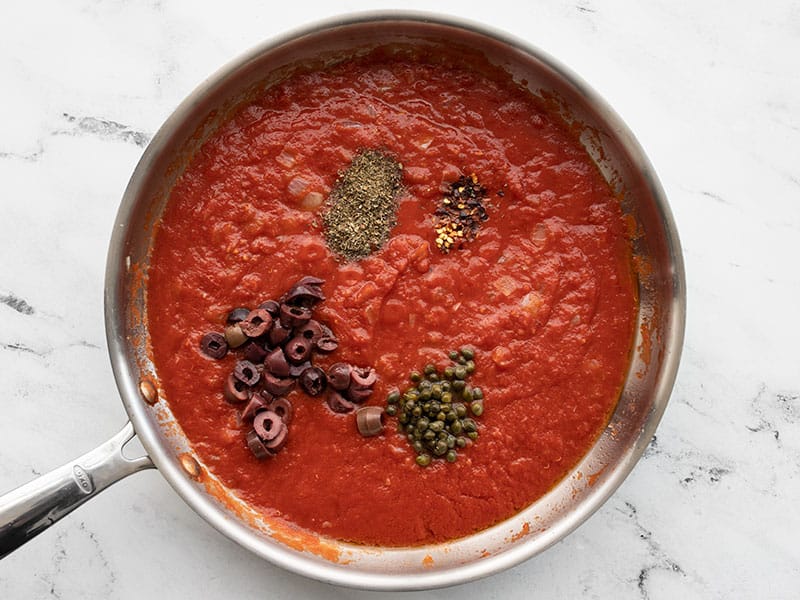 Seasoning the sauce with kalamata olives, capers, basil, and crushed red pepper.
