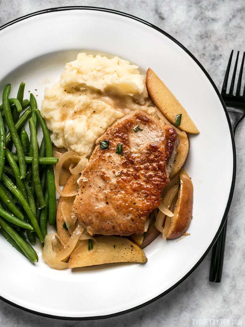These Apple Spice Pork Chops are smothered with apples, onions, cinnamon, and a buttery pan sauce that is sweet, salty, and rich. Budgetbytes.com Apple Spice Pork Chops on a plate with mashed potatoes and green beans