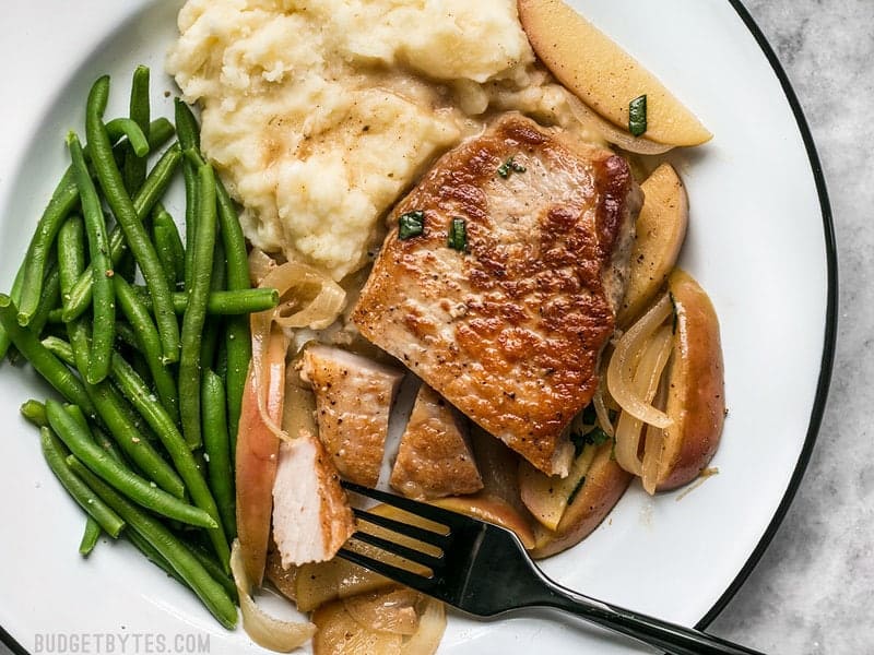 These Apple Spice Pork Chops are smothered with apples, onions, cinnamon, and a buttery pan sauce that is sweet, salty, and rich. Budgetbytes.com Sliced Apple Spice Pork Chops on a white plate with mashed potatoes and green beans.