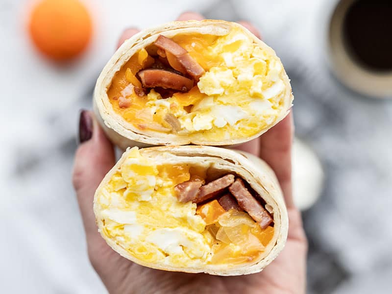 Make ahead breakfast burritos are an easy reheatable and portable breakfast meal prep idea. Including options for vegetarian or other add-ins! BudgetBytes.com The open sides of a cut open breakfast burrito held toward the camera, close up.