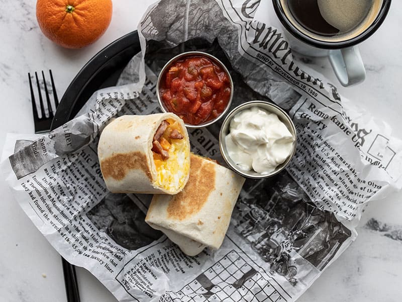 Make ahead breakfast burritos are an easy reheatable and portable breakfast meal prep idea. Including options for vegetarian or other add-ins! BudgetBytes.com A reheated breakfast burrito on a plate with a newsprint liner, two small dishes of salsa and sour cream, a tangerine and cup of coffee on the side