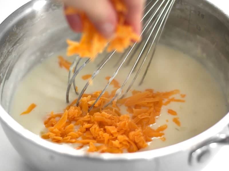 Cheddar cheese being added to pot with cheese sauce