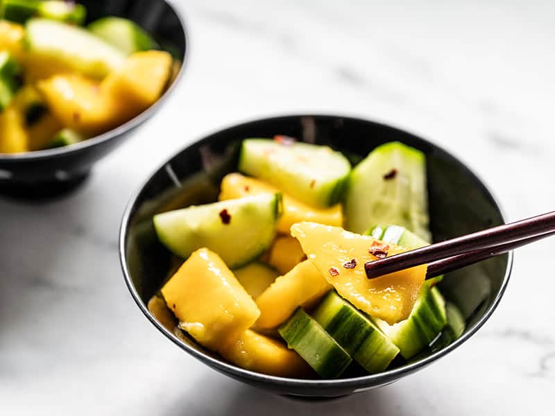 Fresh cucumber and tropical mango come together with a sweet and spicy dressing to make this light and refreshing Cucumber Mango Salad. Budgetbytes.com Close up of a bowl of cucumber mango salad with chopsticks picking up a piece of mango