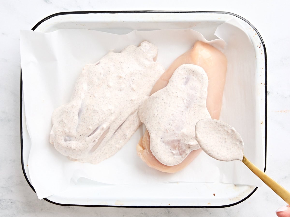 The marinade for chicken shawarma being spooned over two chicken breasts in a parchment lined baking dish.