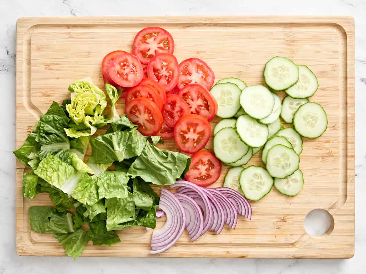 Sliced tomatoes, chopped lettuce, sliced red onion, and sliced cucumber on a wooden cutting board.
