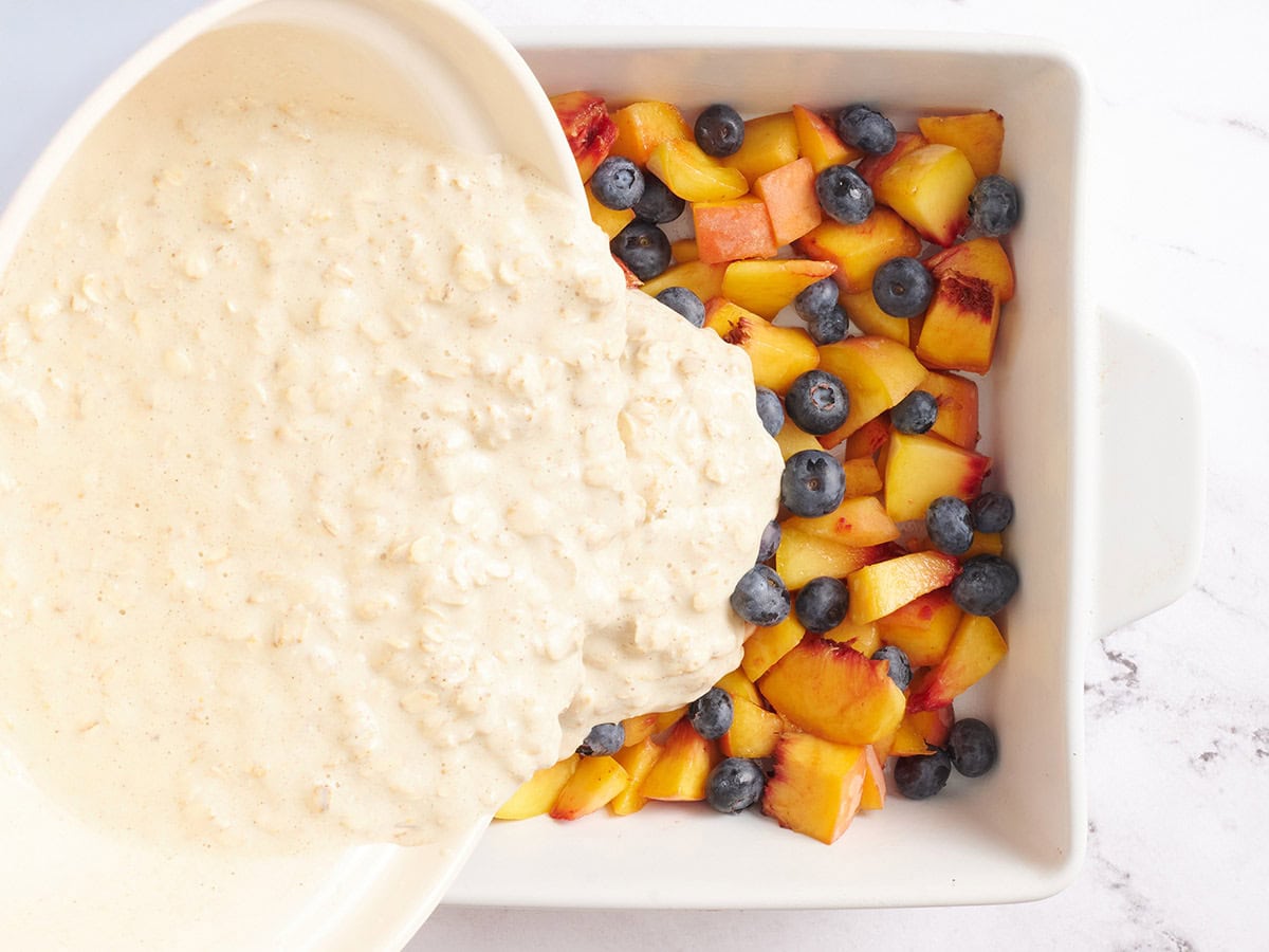 Oatmeal being poured over diced peaches and blueberries in a baking dish.