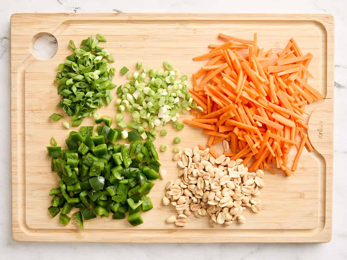 Sliced onion, carrot matchsticks, diced green bell pepper, and peanuts on a wooden cutting board.