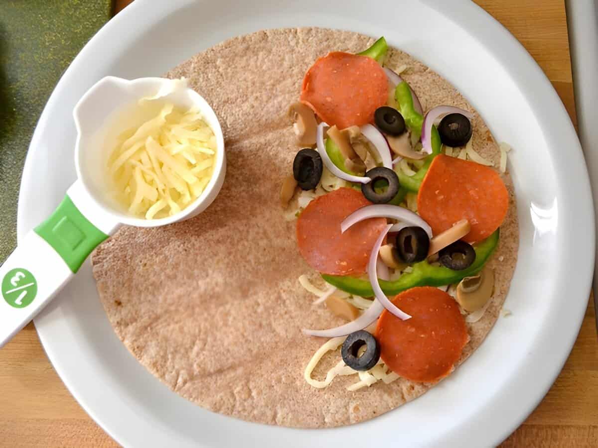 A tortilla topped with shredded cheese, pepperoni, sliced olives, and green bell pepper.