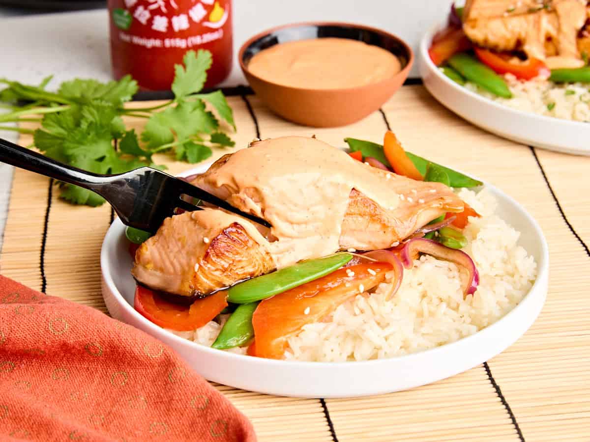 Side view of teriyaki salmon and sriracha mayo on a plate with stir fried vegetables and rice.
