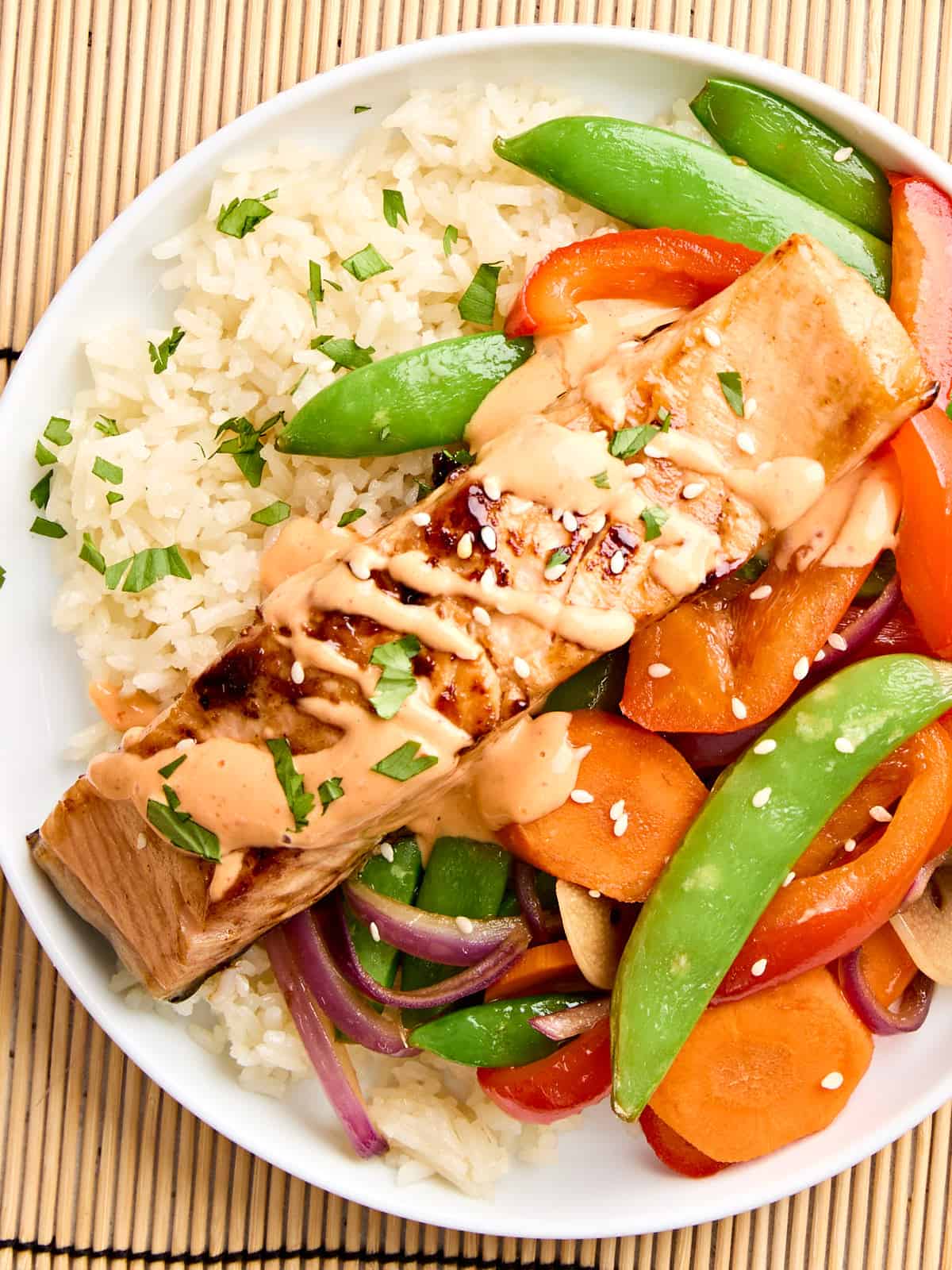 Overhead view of teriyaki salmon and sriracha mayo on a plate with stir fried vegetables and rice.