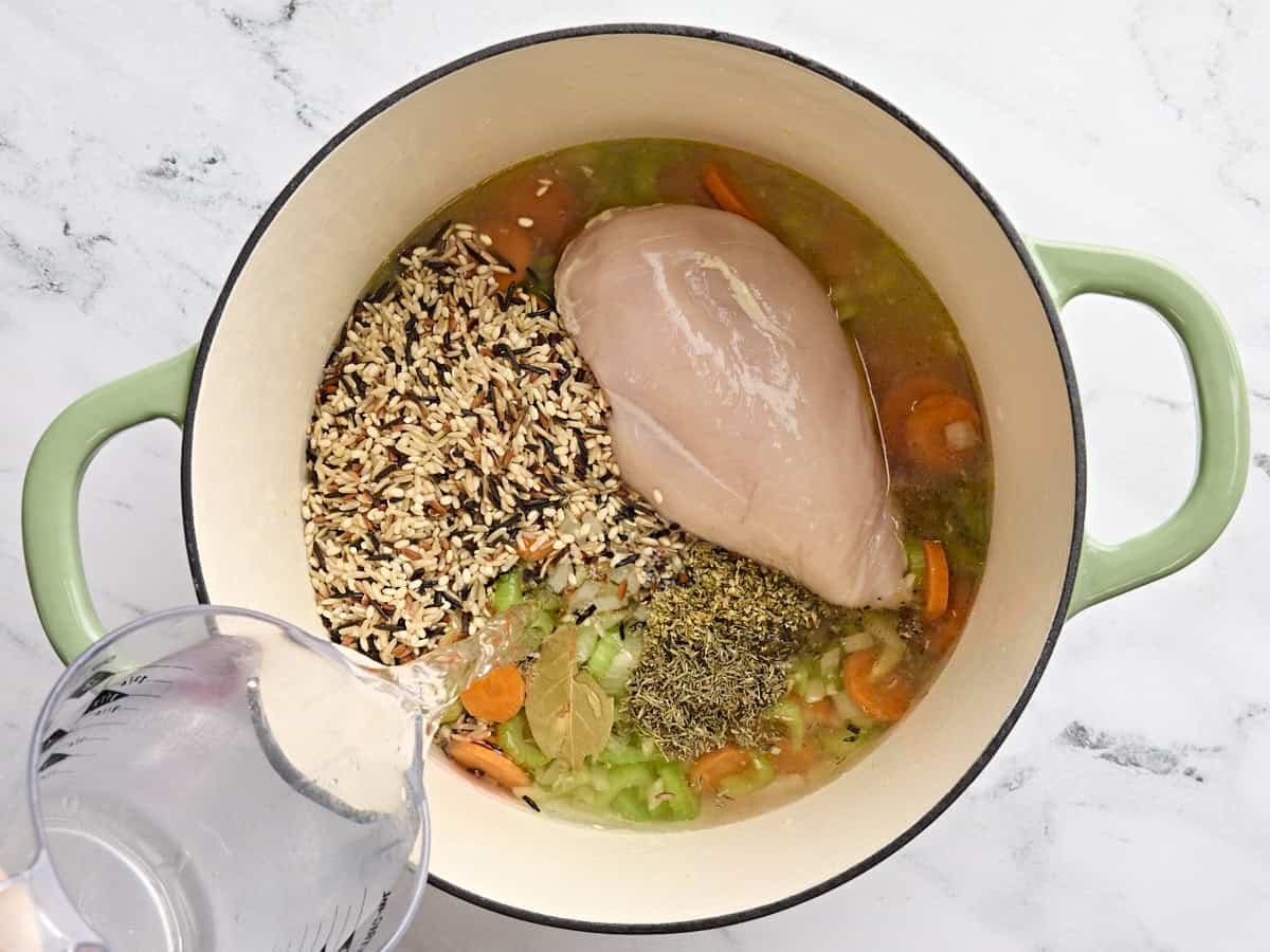 Water being poured into a pot of mirepoix with a chicken breast and wild rice.