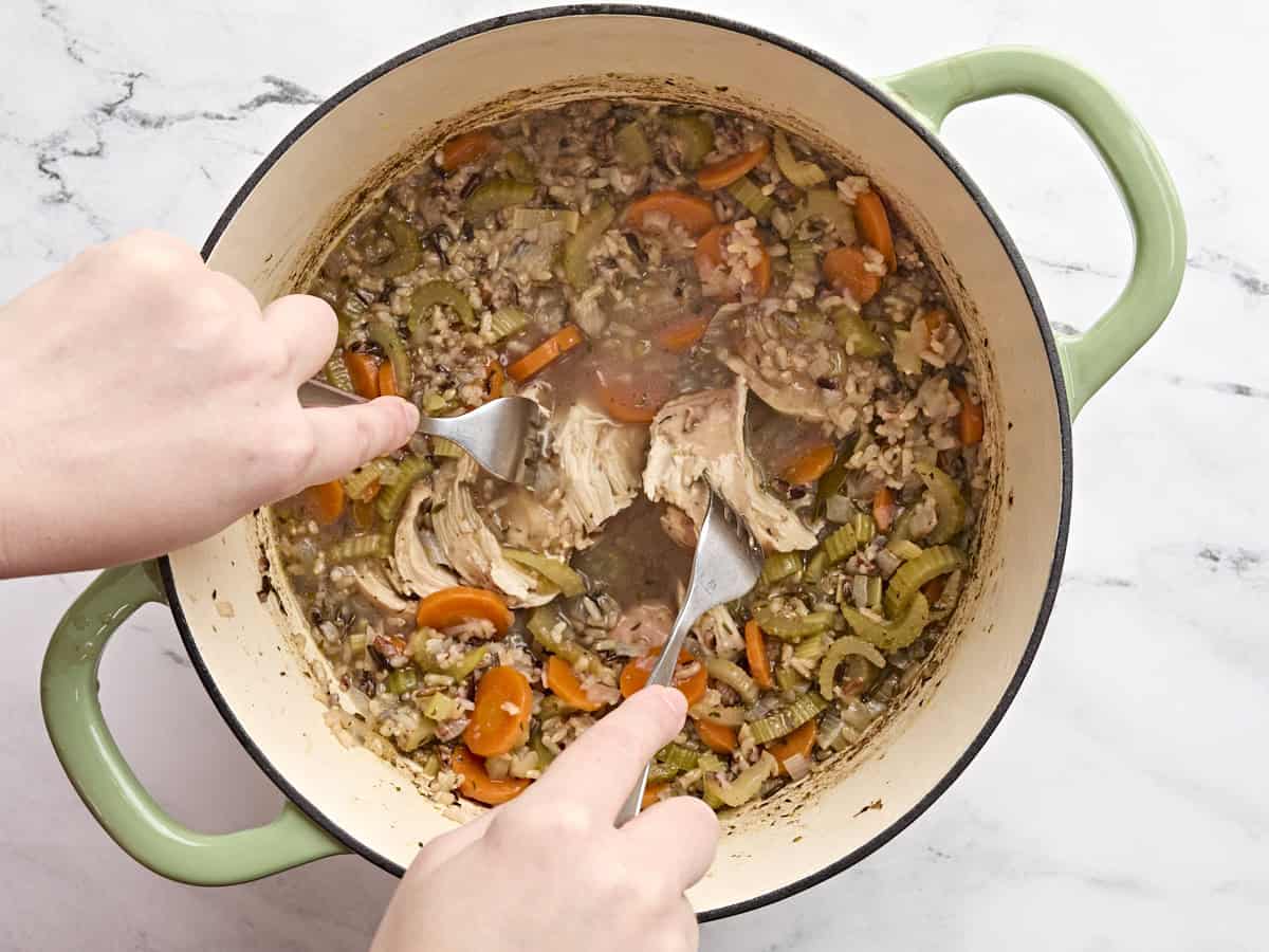 Hands using two forks to shred the chicken in chicken and wild rice soup.