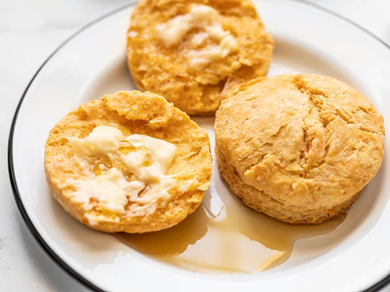 Sweet Potato Biscuits are a unique and subtly sweet twist on a classic, the perfect fall touch for your weekend brunch or Thanksgiving dinner! BudgetBytes.com Close up of two sweet potato biscuits on a plate, one open, smeared with butter, and drizzled with maple syrup