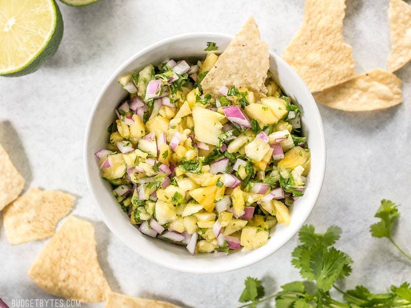 This easy pineapple salsa is fresh, vibrant, and perfect for chips, piling onto grilled meats, topping salads, or adding to any summer meal. BudgetBytes.com A bowl of easy pineapple salsa with chips, lime, and cilantro around the bowl.
