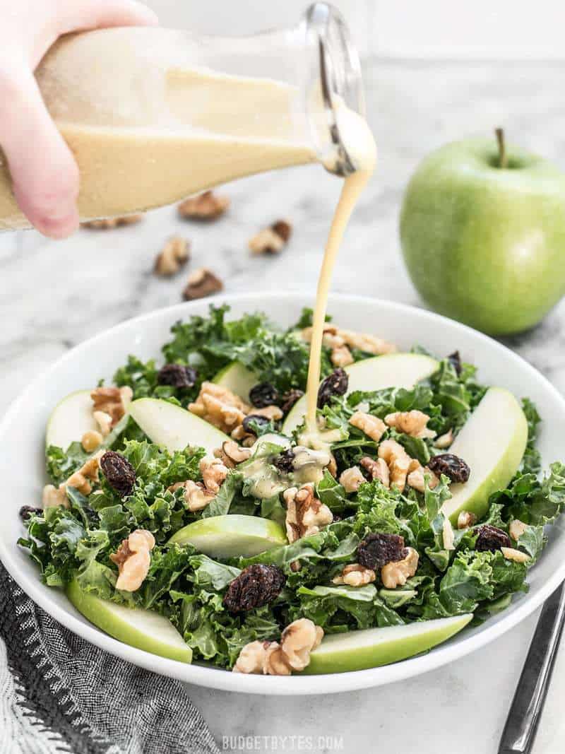 This Apple Dijon Kale Salad is tangy, sweet, and crunchy with Granny Smith apples, walnuts, raisins, and a homemade Dijon vinaigrette. BudgetBytes.com Dijon vinaigrette being poured from a glass bottle onto a bowl of Apple Dijon Kale Salad