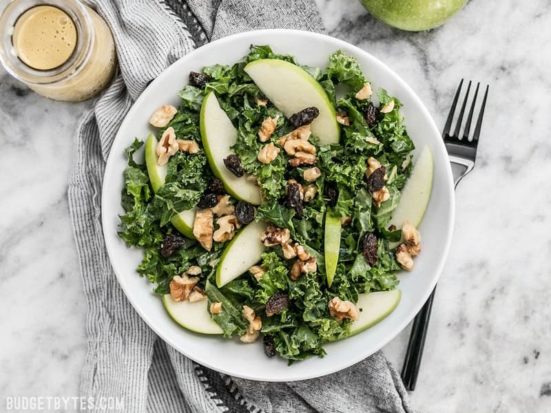 This Apple Dijon Kale Salad is tangy, sweet, and crunchy with Granny Smith apples, walnuts, raisins, and a homemade Dijon vinaigrette. BudgetBytes.com Overhead view of Apple Dijon Kale Salad in a bowl with a black fork