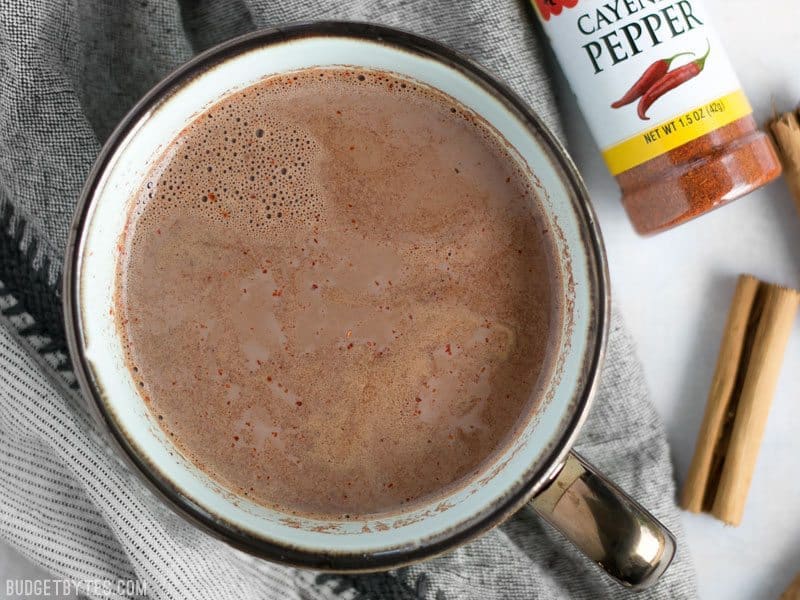 This spicy hot cocoa is rich and warm with a slight kick from cayenne pepper and cinnamon to heat you up inside and out, no matter how cold it is outside! BudgetBytes.com Overhead view of a mug full of Spicy Hot Cocoa with a bottle of cayenne and cinnamon sticks on the side