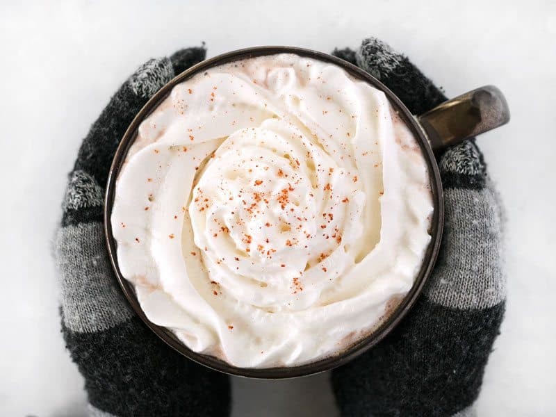 This spicy hot cocoa is rich and warm with a slight kick from cayenne pepper and cinnamon to heat you up inside and out, no matter how cold it is outside! BudgetBytes.com Overhead view of a pair of hands with gloves holding a mug full of spicy hot cocoa topped with whipped cream and a pinch of cayenne