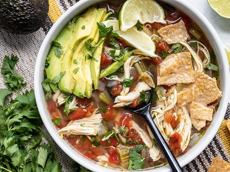 This Chicken and Lime Soup is light, fresh, and flavorful with shredded chicken, vegetables, fresh cilantro, and a tangy lime infused broth. BudgetBytes.com Close up of a bowl of Chicken and Lime Soup with garnishes and a black spoon in the middle