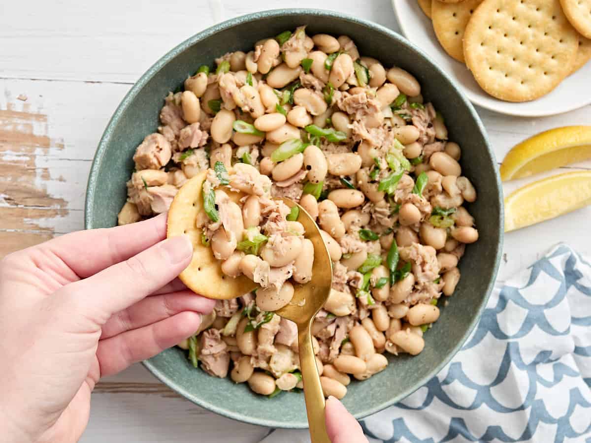 A hand using a spoon to scoop tuna white bean salad onto a cracker,
