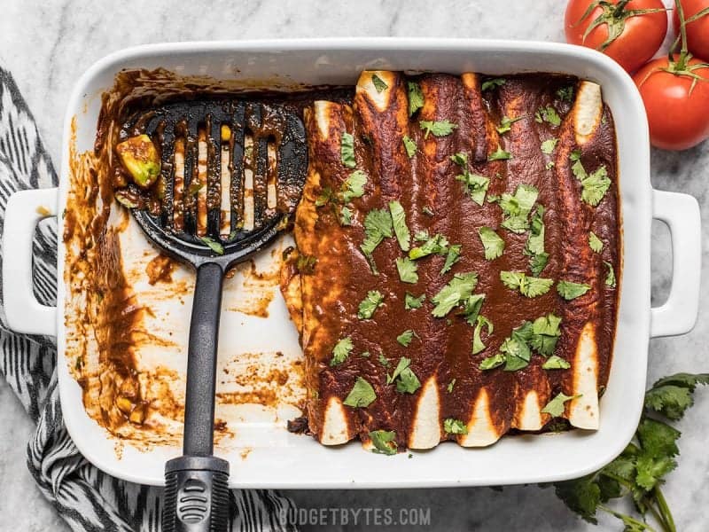 These hearty vegan Black Bean and Avocado Enchiladas are stuffed with fresh ingredients and drenched in a homemade sauce for big flavor in every bite. BudgetBytes.com Casserole dish Black Bean and Avocado Enchiladas, some have been served, spatula resting in their place