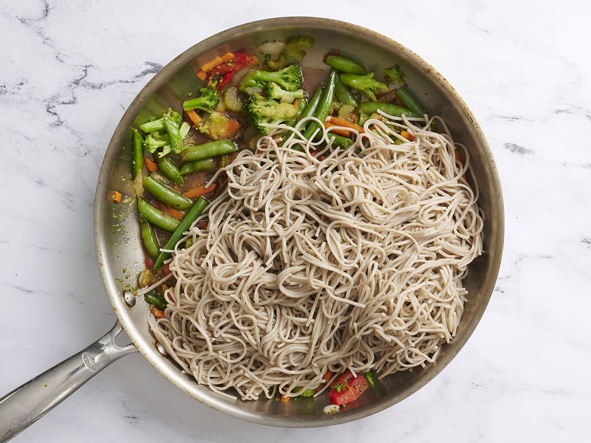 Cooked noodles added to mixed vegetables and teriyaki sauce in a skillet.