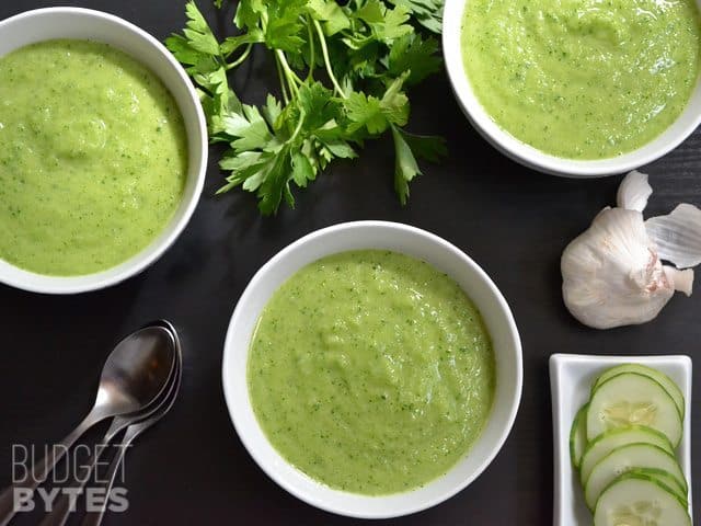 Top view of three bowls of Green Gazpacho with three spoons and a plate of sliced cucumbers