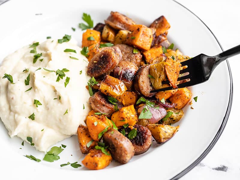 This Oven Roasted Autumn Medley is an herb-infused, sweet and savory sheet pan dinner with sausage, apples, sweet potatoes and more. Budgetbytes.com side view of a fork picking up some oven roasted autumn medley from the plate