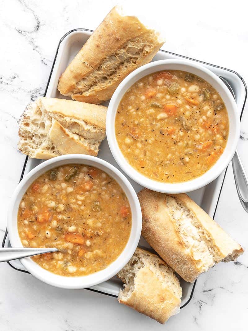 This Slow Cooker White Bean Soup practically makes itself! Just throw everything into the pot and press go for a thick, flavorful, and filling vegan soup. BudgetBytes.com Overhead view of two bowls of slow cooker white bean soup on a white tray with torn pieces of baguette on the sides