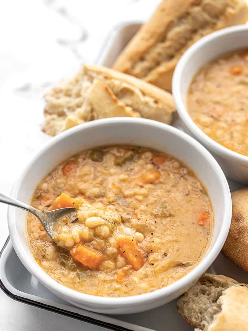 This Slow Cooker White Bean Soup practically makes itself! Just throw everything into the pot and press go for a thick, flavorful, and filling vegan soup. BudgetBytes.com Front view of two bowls of slow cooker white bean soup on a tray, a spoon lifting a bite out of the front bowl