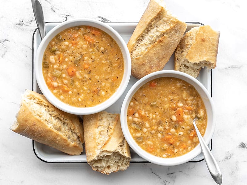 This Slow Cooker White Bean Soup practically makes itself! Just throw everything into the pot and press go for a thick, flavorful, and filling vegan soup. BudgetBytes.com Overhead view of two bowls of slow cooker white bean soup with pieces of baguette on the sides