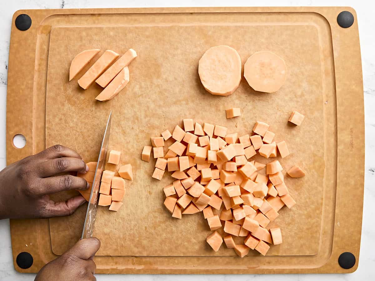 Sweet potatoes being cut into cube on a wooden cutting board.