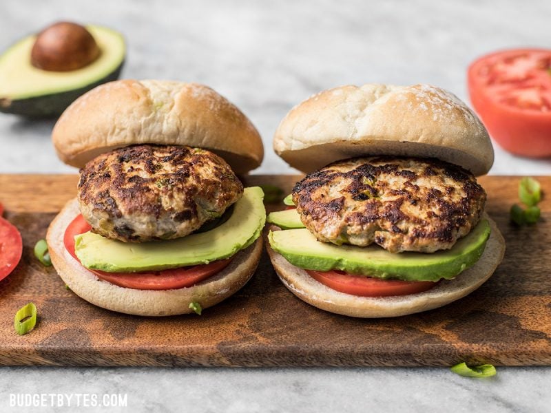 These Green Chile Turkey Burgers can't be beat for a fast, flavorful weeknight dinner. Healthy, ready in about 30 minutes, and full of southwest flavor. Budgetbytes.com Side view of two Green Chile Turkey Burgeres with tomato and avocado in the background.