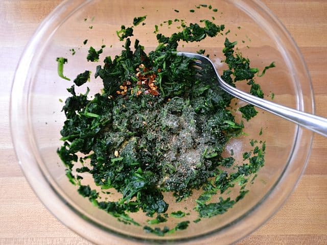 Seasoning mixed into frozen spinach in mixing bowl with fork