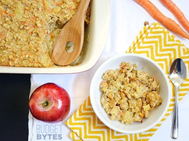 Top view of a bowl of Morning Glory Baked Oatmeal with full pan of oatmeal, an apple and a spoon on the side