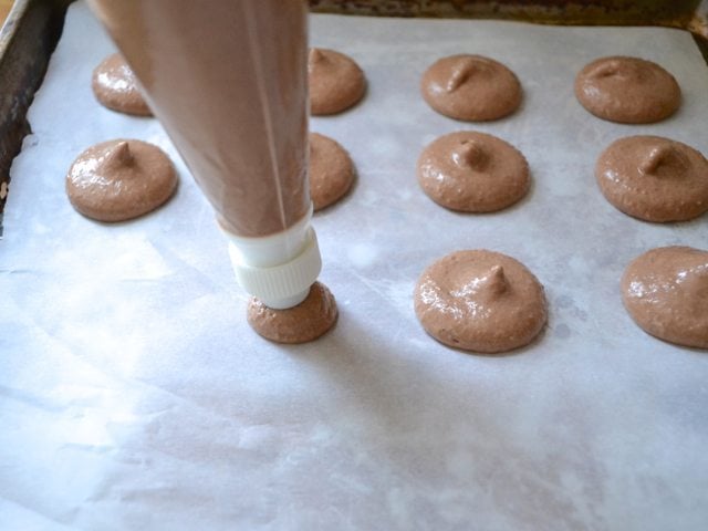 Piping macarons out of pastry bag onto baking sheet lined with parchment paper