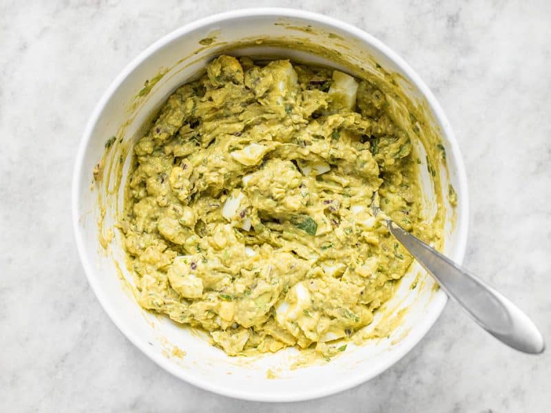 Chopped eggs stirred into other ingredients in mixing bowl with spoon