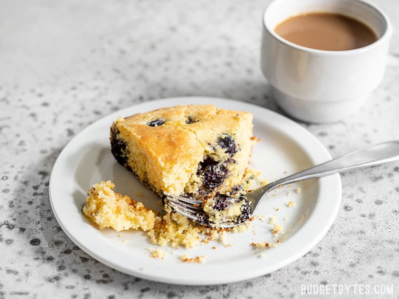 For a deliciously lazy weekend treat, whip up this super fast and easy Lemon Blueberry Cornbread Skillet. Great for breakfast, dessert, or coffee! Budgetbytes.com A slice of Lemon Blueberry Cornbread on a plate with a fork and cup of coffee in the background.