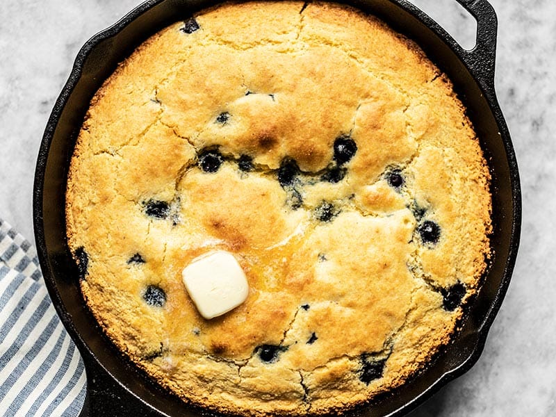 For a deliciously lazy weekend treat, whip up this super fast and easy Lemon Blueberry Cornbread Skillet. Great for breakfast, dessert, or coffee! Budgetbytes.com Close up of baked Lemon Blueberry Cornbread in the skillet with some butter melting on top.