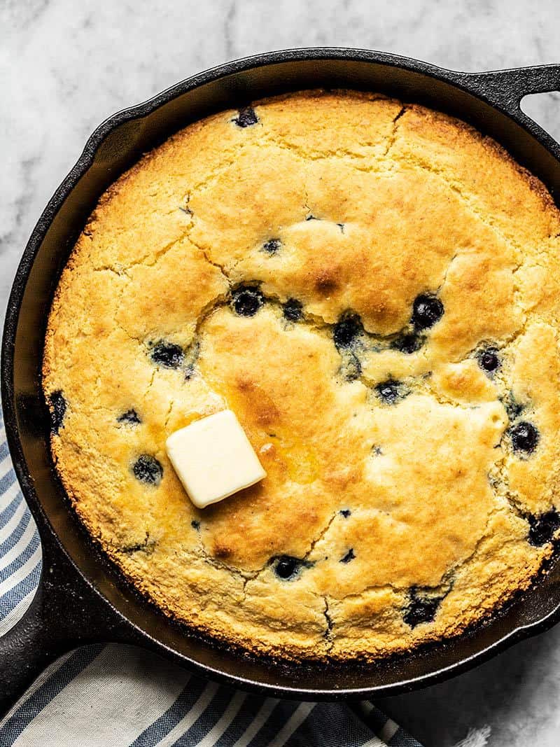 For a deliciously lazy weekend treat, whip up this super fast and easy Lemon Blueberry Cornbread Skillet. Great for breakfast, dessert, or coffee! Budgetbytes.com Close up of baked Lemon Blueberry Cornbread in a cast iron skillet with a pat of butter melting on top.