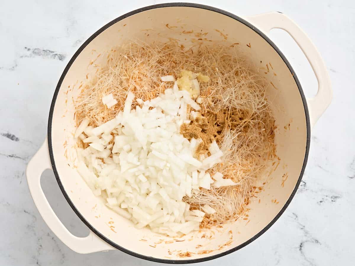 Diced onion, cumin, and minced garlic added to toasted noodles in a pot.