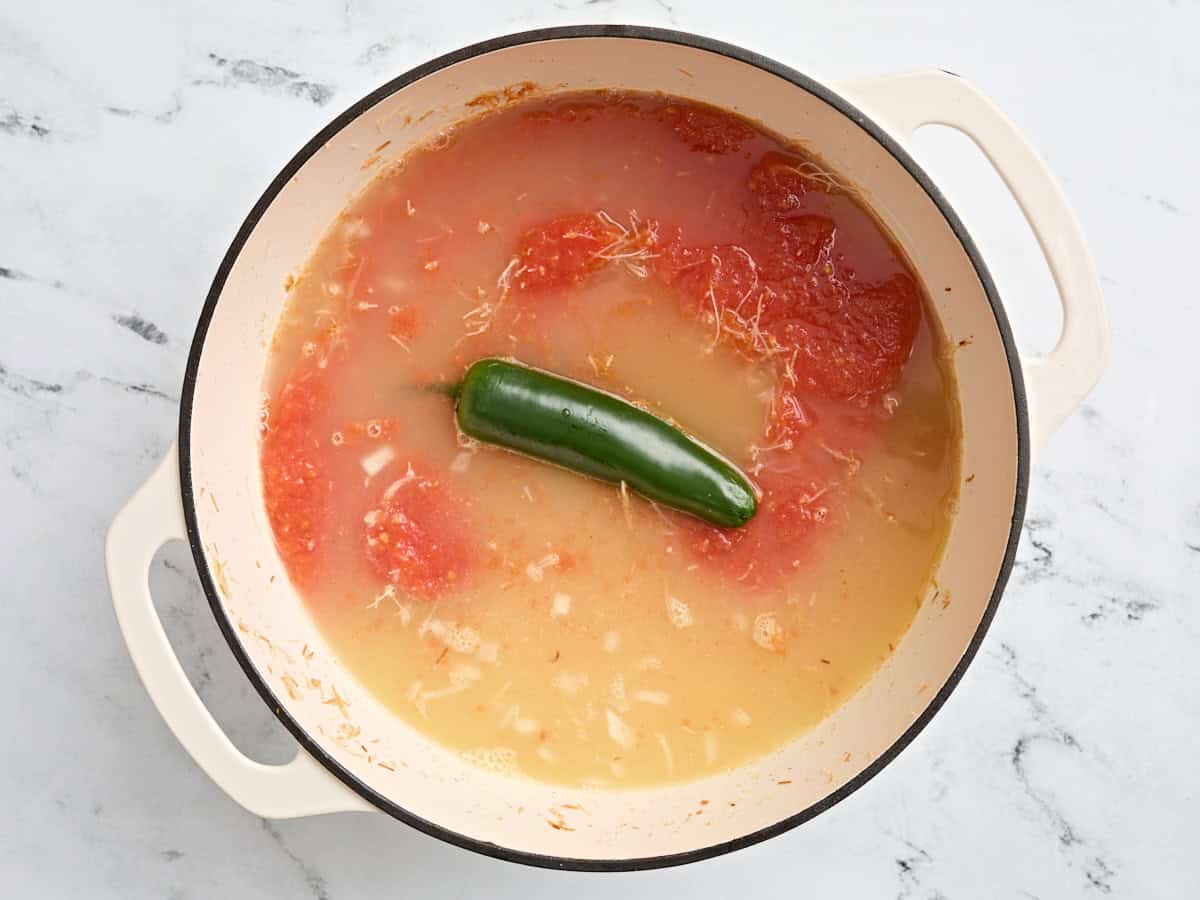 Chicken broth, a whole jalapeno, and pureed tomatoes in a pot with toasted noodles and onions.