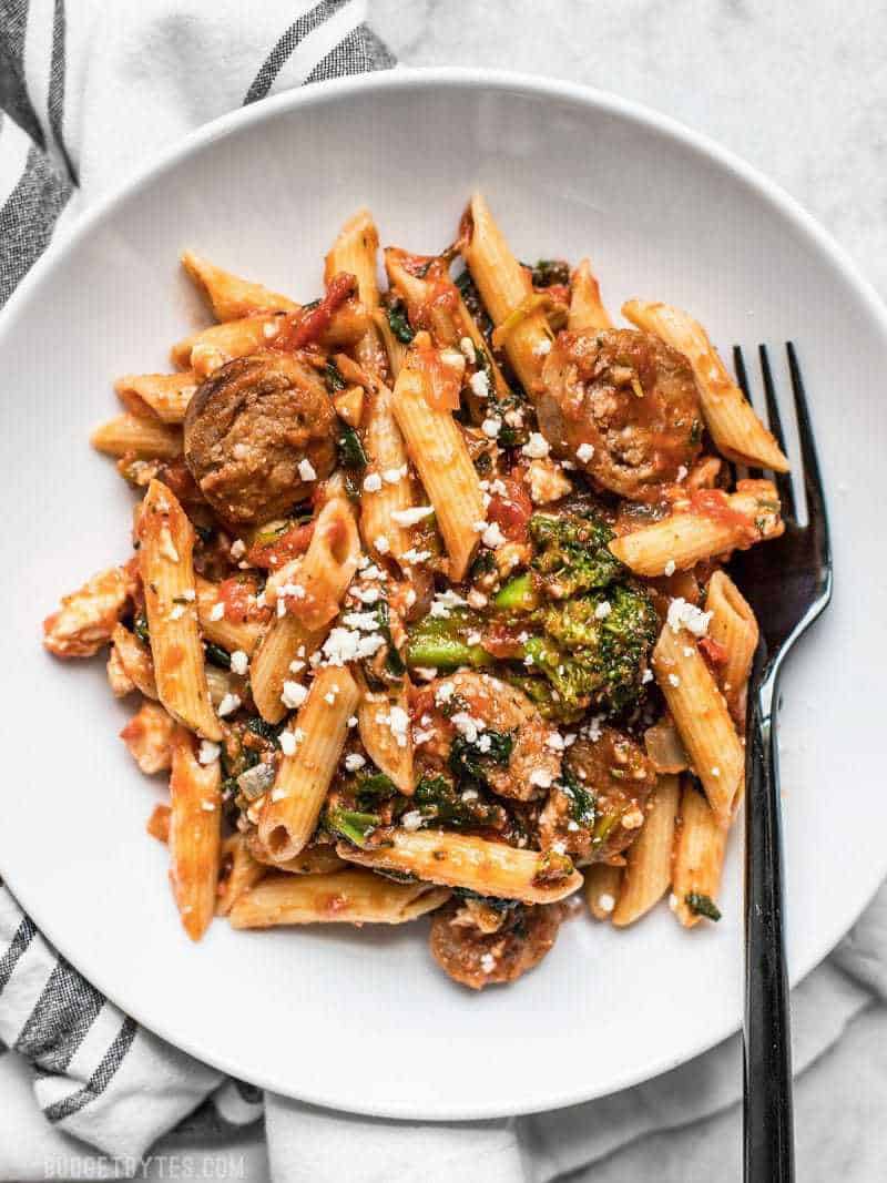 This super easy and satisfying, "sweep the kitchen" Penne Pasta with Sausage and Greens is an entire meal in one pot. Cook once, eat all week. A white plate full of Penne Pasta with Sausage and Greens, and a black fork.